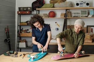 Teenage guy and his grandmother with wrenches fixing wheels of skateboard while standing by table with handtools in garage