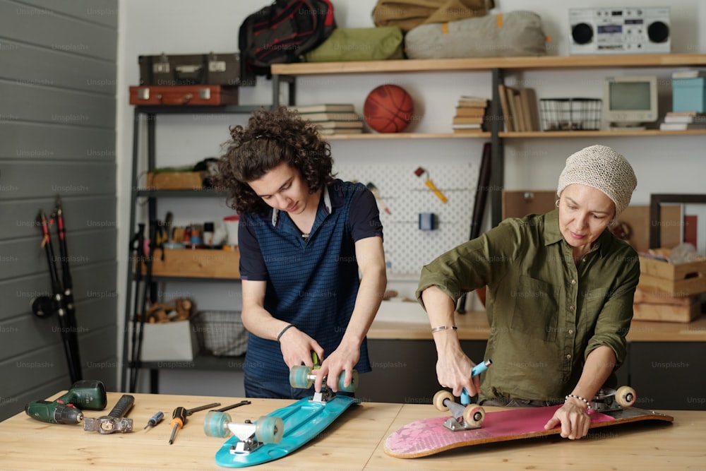 Teenage guy and his grandmother with wrenches fixing wheels of skateboard while standing by table with handtools in garage