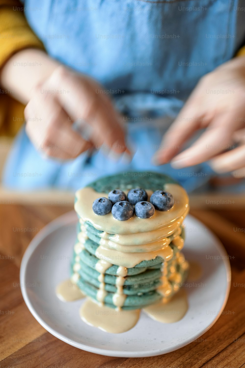 青いエプロンを着た女性は、木製のテーブルにキャラメルのスーズを注いだブルーベリーで青いアメリカのパンケーキを飾ります。自家製のおいしい料理。シュローブタイドの祭典。