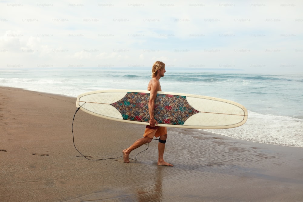 サーファー。砂浜を歩く白いサーフボードを持つハンサムなサーフィンマン。美しい海を背景にアクティブなライフスタイルのためのウォータースポーツ。