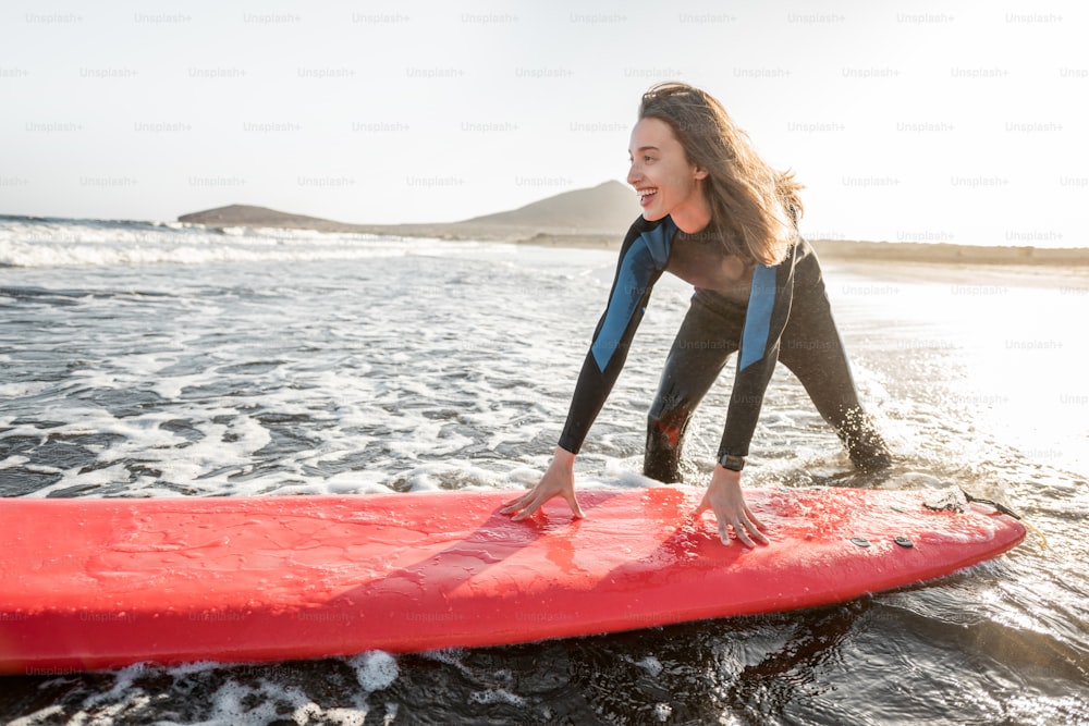 Joven surfista en traje de neopreno que se sube a la tabla de surf, atrapando el flujo de agua cerca de la playa durante una puesta de sol. Concepto de deportes acuáticos y estilo de vida activo