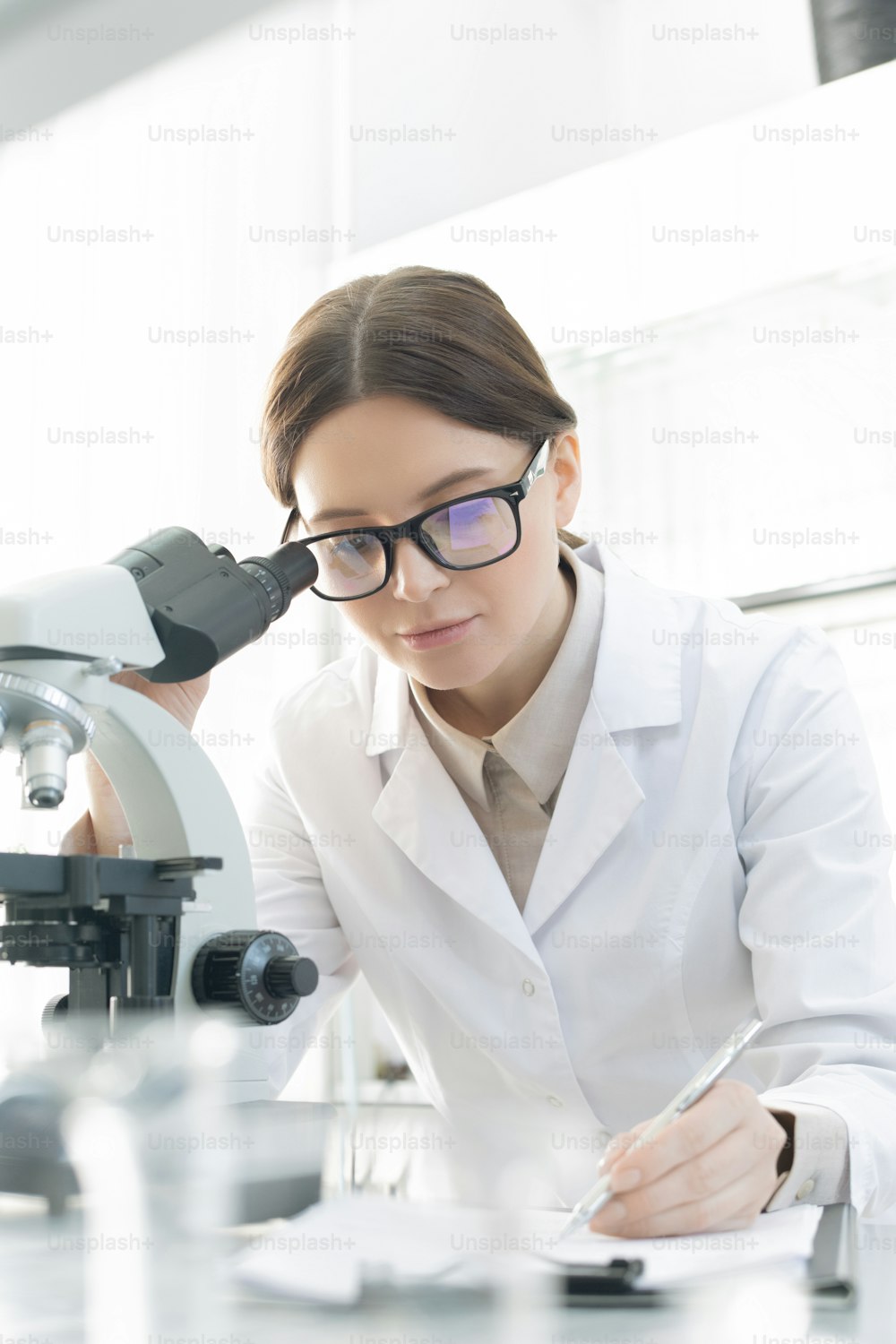 Brunette female scientific researcher in whitecoat making notes while studying coronavirus characteristics in microscope in lab