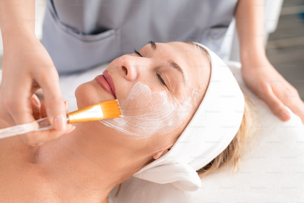 Close-up of beauty professional applying facial mask to relaxed mature woman with closed eyes at spa procedure