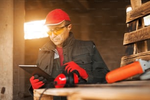 Modern senior carpenter in a professional uniform looking on a tablet while standing near electric tool on a wood pallet.