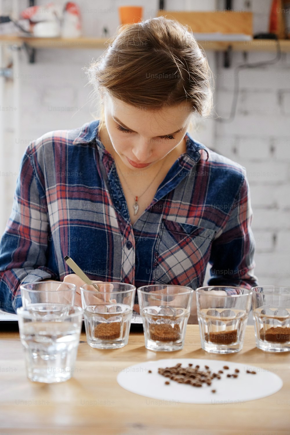 Retrato da jovem barista feminina que escreve o resultado do teste de xícara de café, examinando xícaras com grãos na mesa de madeira