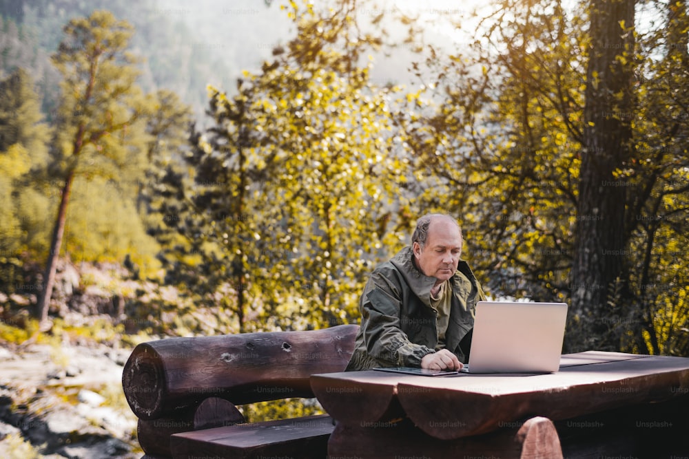 Mature man in overalls is working on laptop while sitting at the wooden table in parkland; hunter forester is sitting on the bench and using his netbook with hills and wood in the blurry background