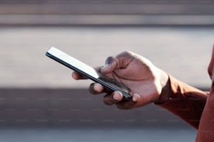 Mano de un joven africano con un teléfono inteligente enviando mensajes de texto o desplazándose a través de cosas en línea al aire libre