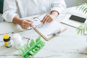 여성 영양사 의사의 자르기 사진 플러스 크기 흰색 셔츠는 밝고 현대적인 사무실에서 매일 알약과 메모가 있는 테이블에서 일하고 있습니다.