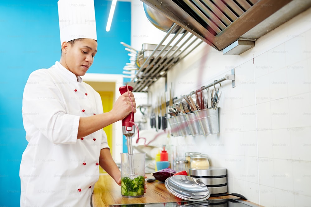 Young woman in chef uniform making broccoli smoothie with electric blender