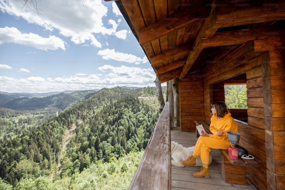 Young woman enjoys great mountain landscape and prepares food, while sitting with her dog on a wooden terrace of nature house. Concept of escape and solitude in nature and traveling with pets
