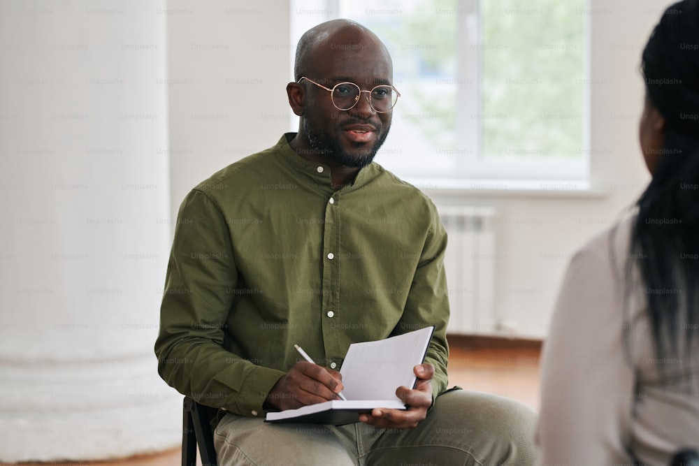 Jeune homme confiant prenant des notes dans un cahier lors d’une séance pendant une conversation avec une patiente assistant à un cours d’aide psychologique