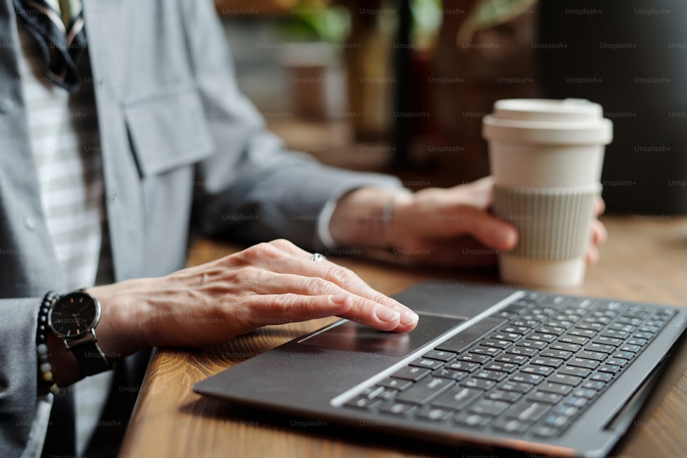 Hand einer jungen Geschäftsfrau oder Freiberuflerin über Laptop-Tastatur, die im Café sitzt und Kaffee trinkt, während sie an einem neuen Projekt arbeitet