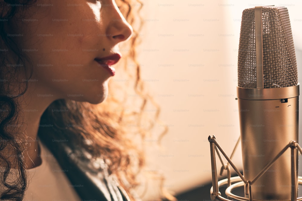 Close up of beautiful curly woman in leather jacket and hat with red lips recording vocals in music studio on professional sound equipment