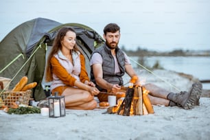 Young and cheerful couple sitting at the fireplace, cooking sausages, having a picnic at the campsite on the beach in the evening