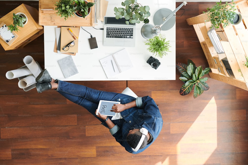 Young millennial in denim casualwear sitting on chair by desk while using paint program in tablet and working over design project