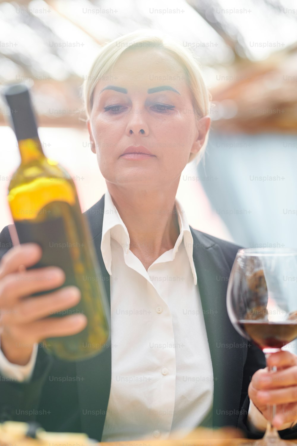 Mature blonde woman in formalwear looking at bottle of cabernet while estimating quality of wine