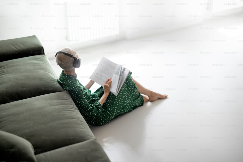 Femme élégante en robe verte lisant un livre et écoutant de la musique, assise sur le sol près du canapé à la maison. Loisirs et confort de vie à la maison