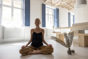 Junge stilvolle Frau, die in einem modernen, hellen Wohnzimmer zu Hause meditiert und Yoga praktiziert. Achtsamkeit, Ruhe im modernen Zuhause
