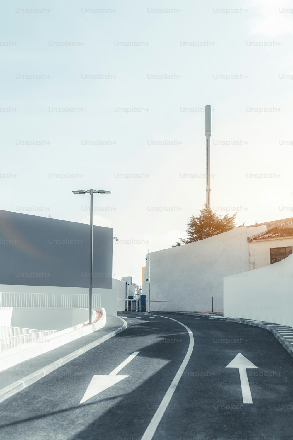 Una vista vertical de una estrecha carretera asfaltada de dos carriles que sigue a la barrera al final con flechas opuestas como marca vial y pequeñas aceras a ambos lados, la antena celular detrás, día soleado