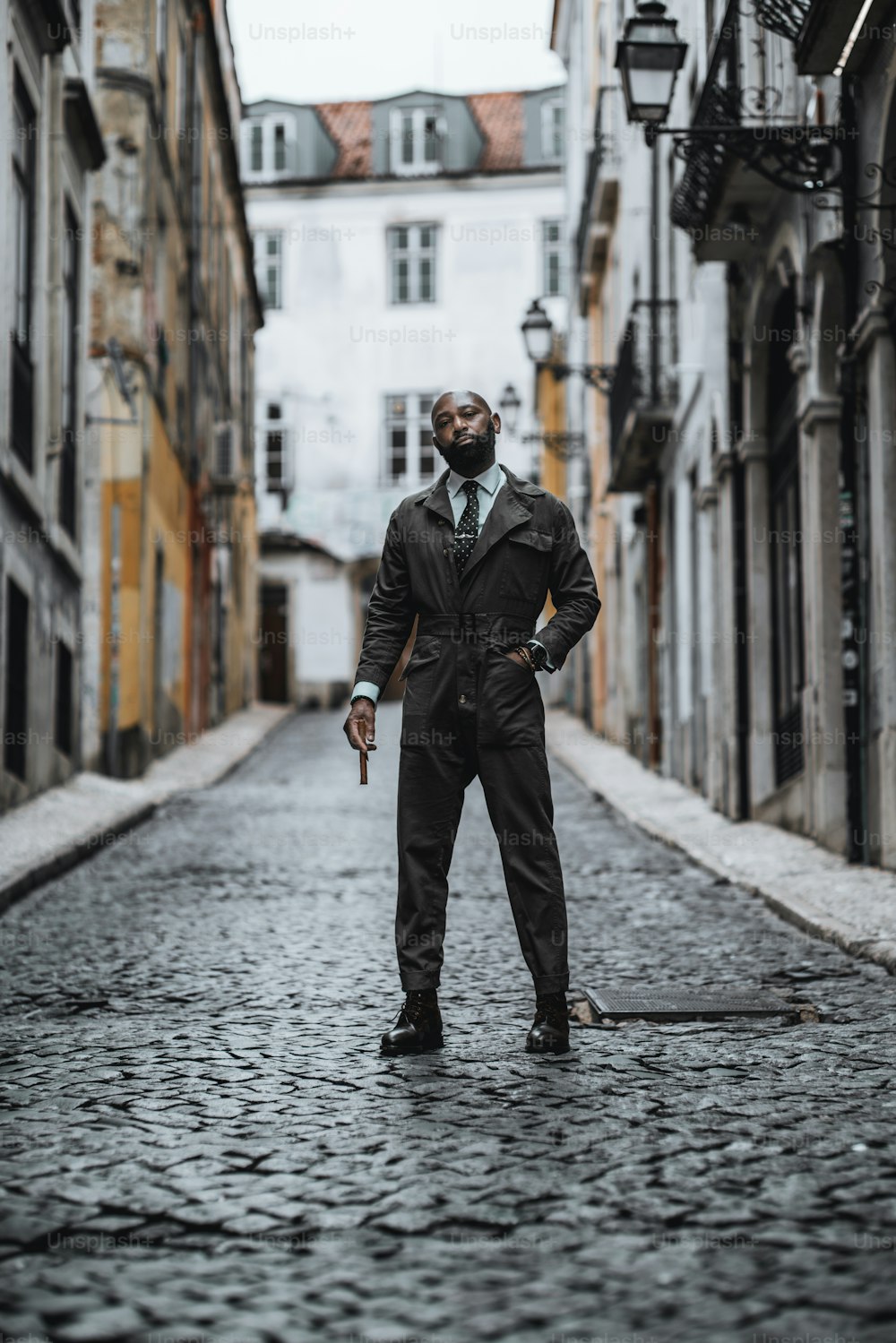A fashionable mature bearded bald African-American man in dark-green elegant custom-made overalls is standing on a paving stone of a narrow European street with a cigar in his hand; selective focus