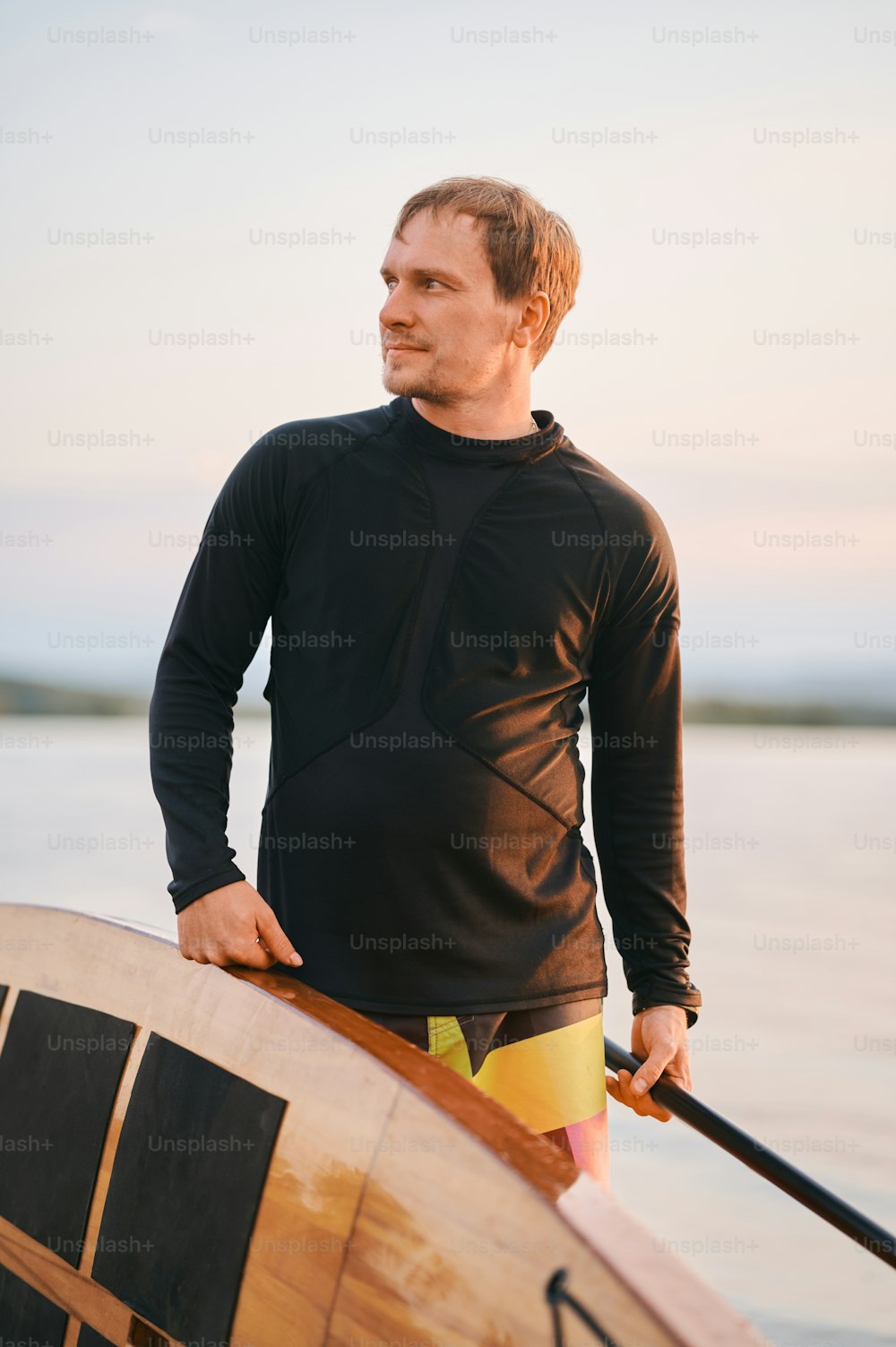 Portrait d’un homme fort en chemise mouillée posant avec une planche de surf et une pagaie au bord du lac après avoir pagayé activement sur l’eau, en regardant loin de la caméra