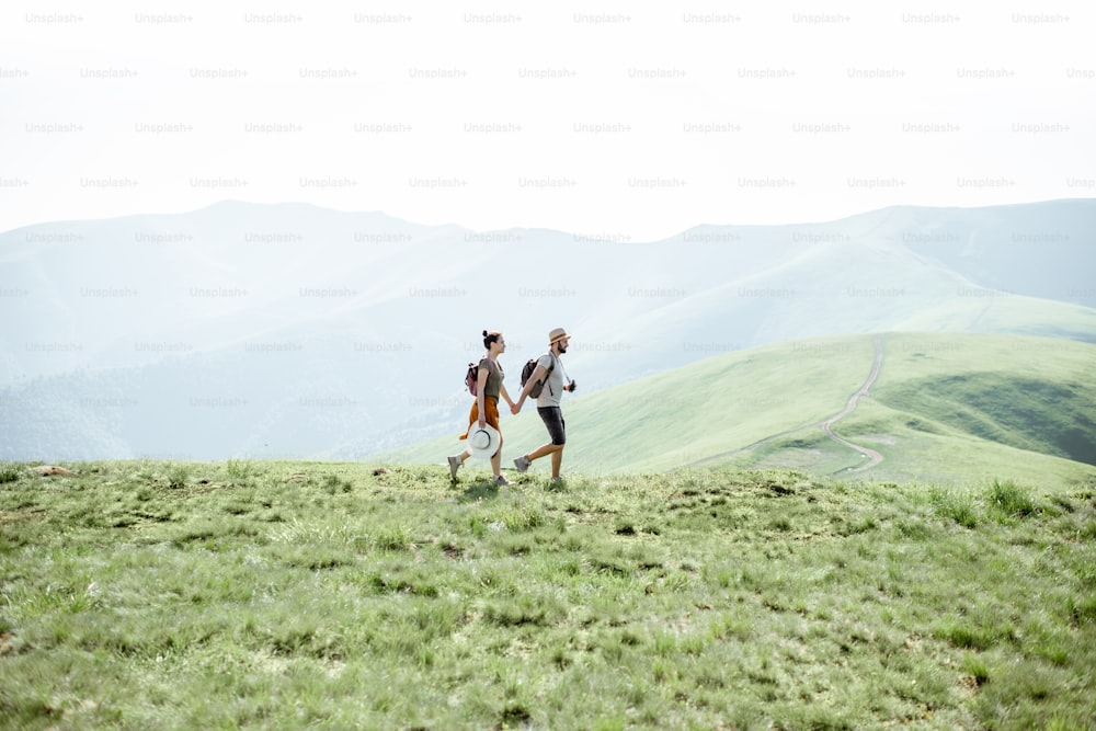 Couple walking with backpacks on the green meadow, traveling in the mountains during the summer time, wide landscape view