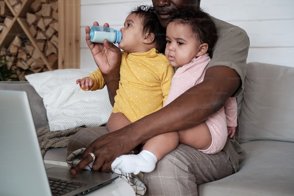 Modern African American father sitting on sofa in living room working on laptop and taking care of his baby daughters at the same time
