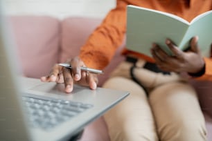 Mains d’une jeune femme africaine avec un stylo touchant le panneau par clavier tout en étant assise devant un ordinateur portable et en apprenant