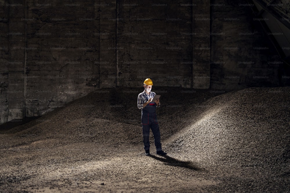 Quality control of semi-raw materials in the industry. A senior quality controller with a helmet on his head standing in a factory hall full of sugar beet products and using a tablet.