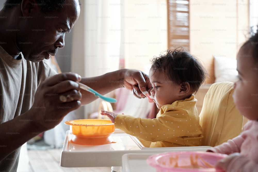 현대의 성숙한 아프리카 계 미국인 남자는 맛있는 과일 퓌레로 먹이는 쌍둥이 딸을 돌보고 있습니다.