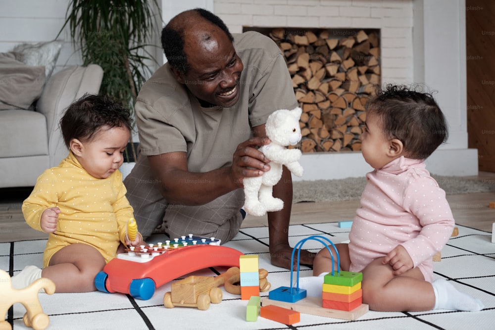 행복한 아프리카계 미국인 아버지는 집 거실 바닥에 앉아 귀여운 쌍둥이 아기들과 함께 부드러운 장난감을 보여주고 놀고 있습니다.
