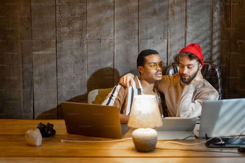 Two men with different nationality having fun while working on computers at home office. Concept of gay couples working together. Caucasian and hispanic man together