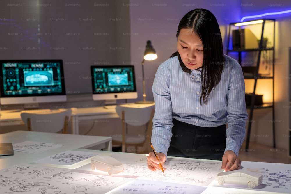 Konzentrierte junge asiatische Autodesignerin steht am Schreibtisch und zeichnet im dunklen Büro eine Auto-Außenskizze