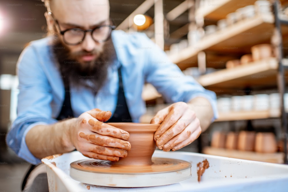 Hombre guapo como trabajador de alfarero en delantal haciendo jarras de arcilla en la rueda de cerámica en la pequeña fábrica