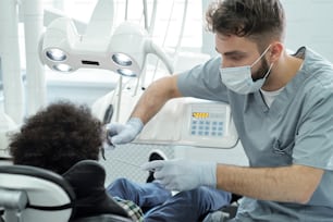 Dentista de uniforme, máscara e luvas segurando broca dentária e instrumento por via oral de pequeno paciente enquanto se inclina sobre ele