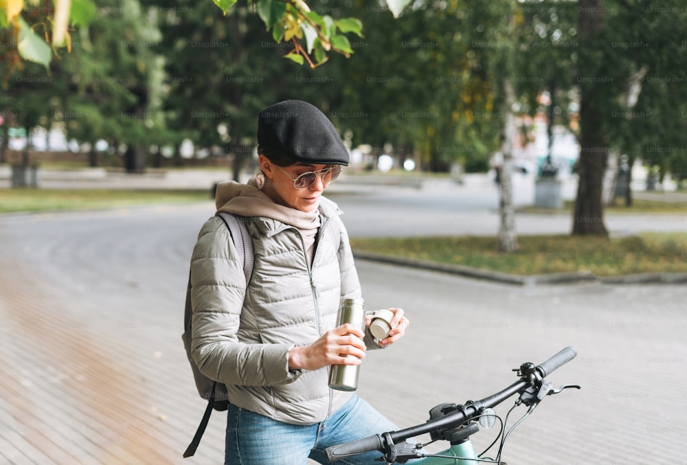 Portrait of fashionable young pretty woman in cap and sunglasses on bicycle with thermo mug on sunny autumn day in city