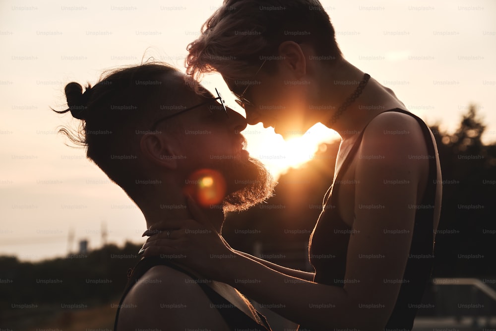 Primo piano ritratto di giovane coppia in occhiali rotondi con capelli colorati che si baciano all'aperto nella luce naturale del tramonto. Il raggio di sole splende tra di loro