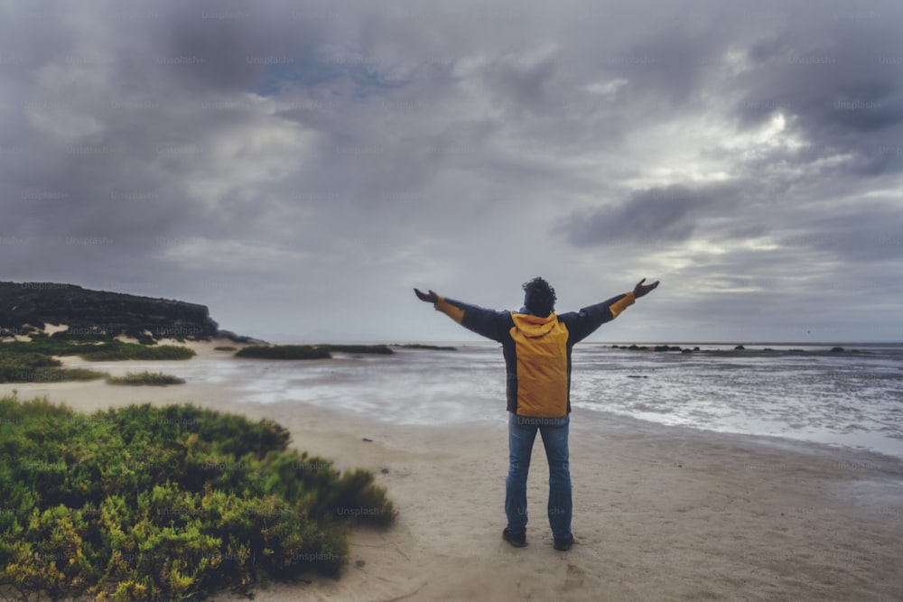 Travel explore destination with one man viewed frmo back outstretching arms for satisfaction and happiness in fron tof a wild bad weather beach and dramatic sky with ocean