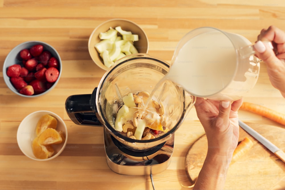 Hands of female pouring milk into electric blender with chopped ingredients while preparing smoothie