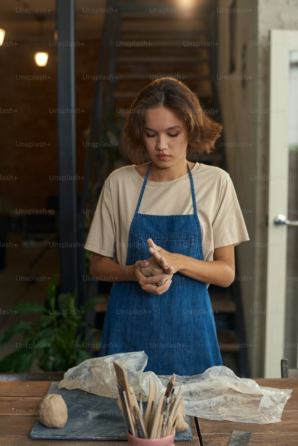 Young craftswoman in workwear kneading piece of clay between hands while preparing it for further processing