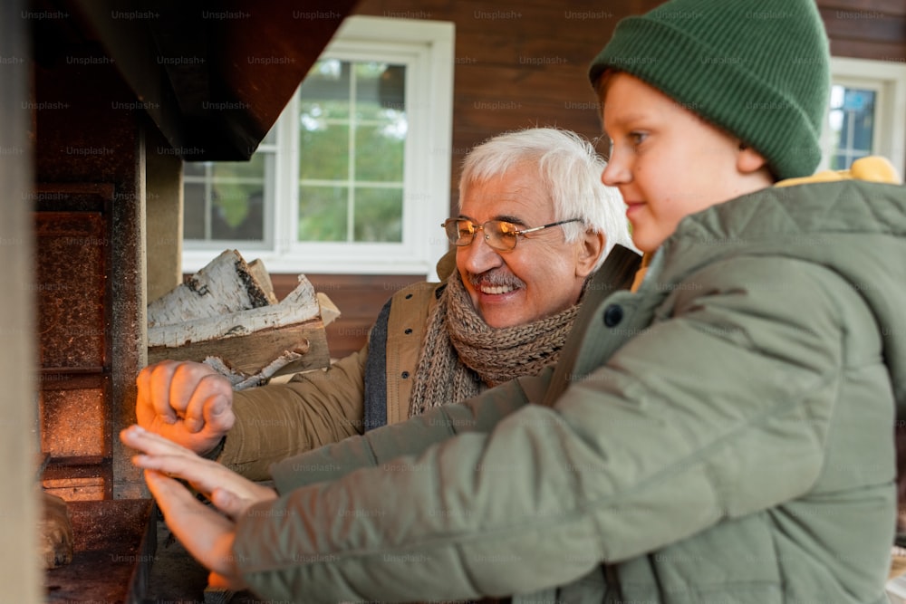 Happy senior man and his grandson standing by fireplace with burning firewoods while getting warm on cool autumn day by country house