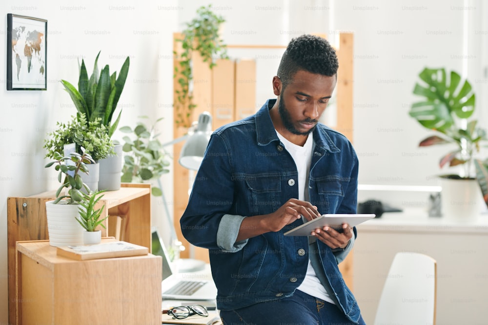 Jovem hipster sério em jaqueta jeans rolando na guia digital deixar enquanto navega para dados on-line ou sites de design no escritório