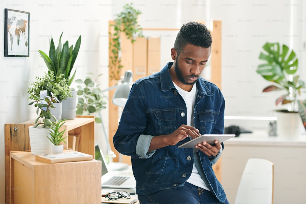 Young serious hipster man in denim jacket scrolling in digital tab let while surfing for online data or design websites in office