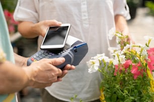 Joven cliente de floristería o centro de jardinería sosteniendo un teléfono inteligente sobre la máquina de pago mientras compra flores