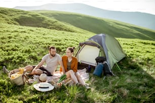 Young and cheerful couple having a picnic at the campsite while traveling high in the mountains during the sunset