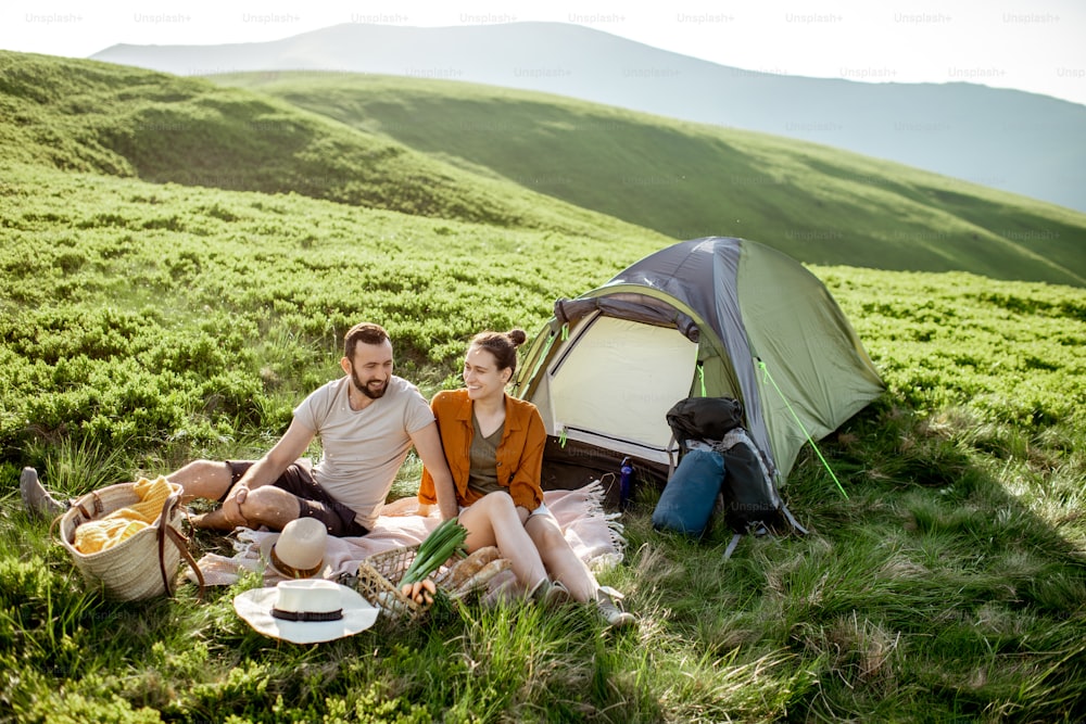 Pareja joven y alegre haciendo un picnic en el camping mientras viaja por lo alto de las montañas durante la puesta de sol