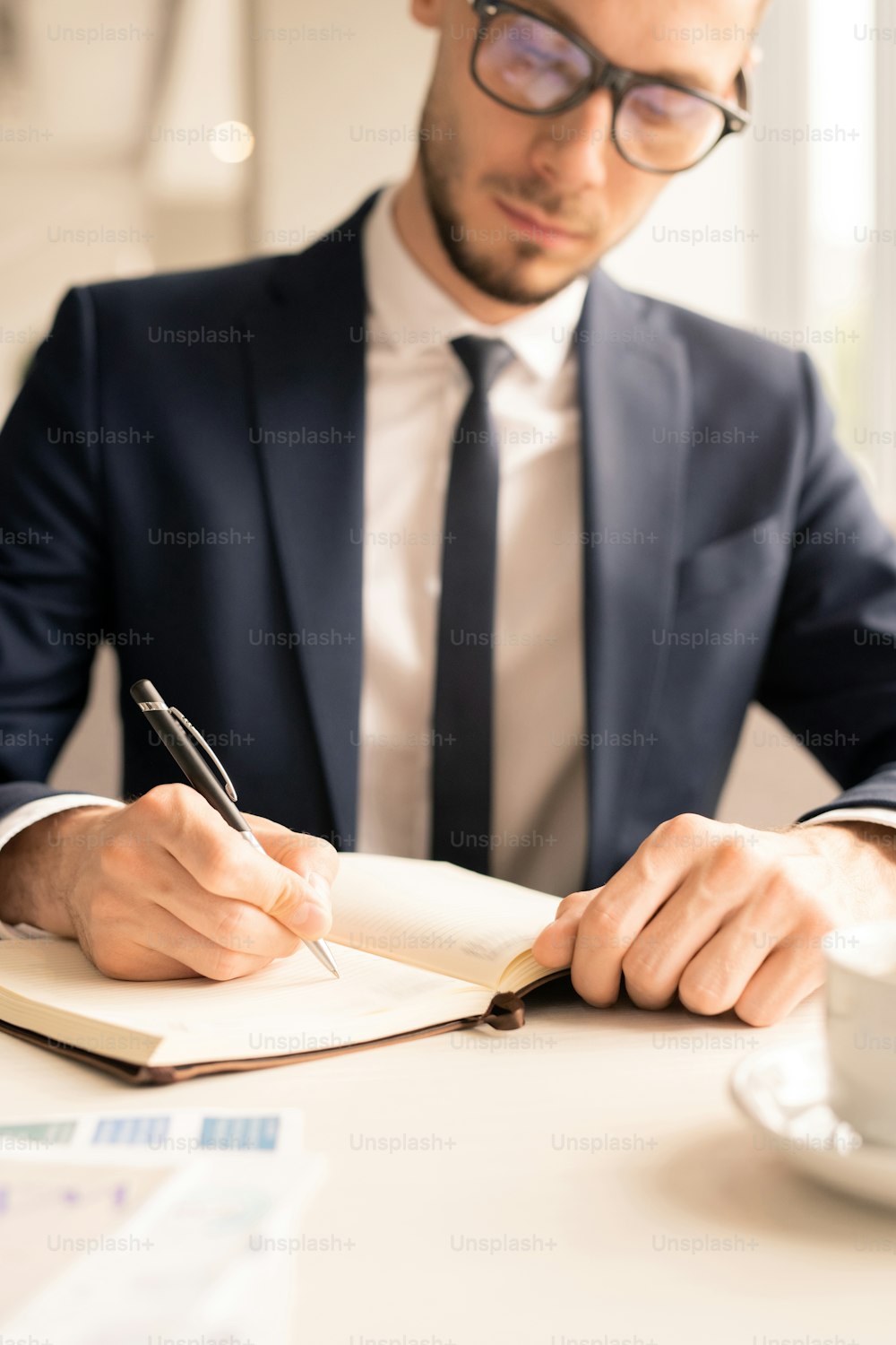 Empleado joven serio en traje sentado junto a la mesa frente al cuaderno abierto y tomando notas o puntos del plan de negocios