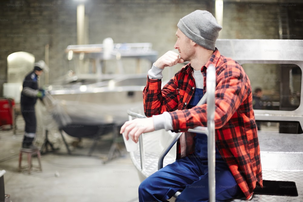 Young engineer sitting on metallic boat and looking at his colleague working in factory