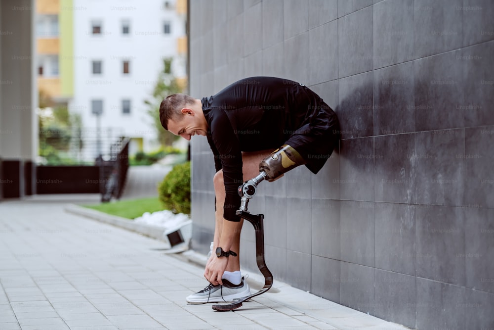 Vista laterale di attraente sportivo caucasico con gamba artificiale appoggiata al muro e allacciandosi i lacci delle scarpe.