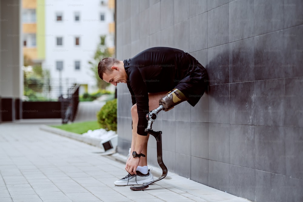 Vista lateral de un atractivo deportista caucásico con una pierna artificial apoyada en la pared y atándose el cordón de los zapatos.
